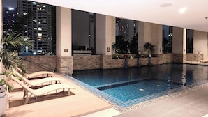 A modern indoor swimming pool with a tiled floor, lounge chairs, and a view of a cityscape at night. - KL Executive Greenbelt KLX (Makati)