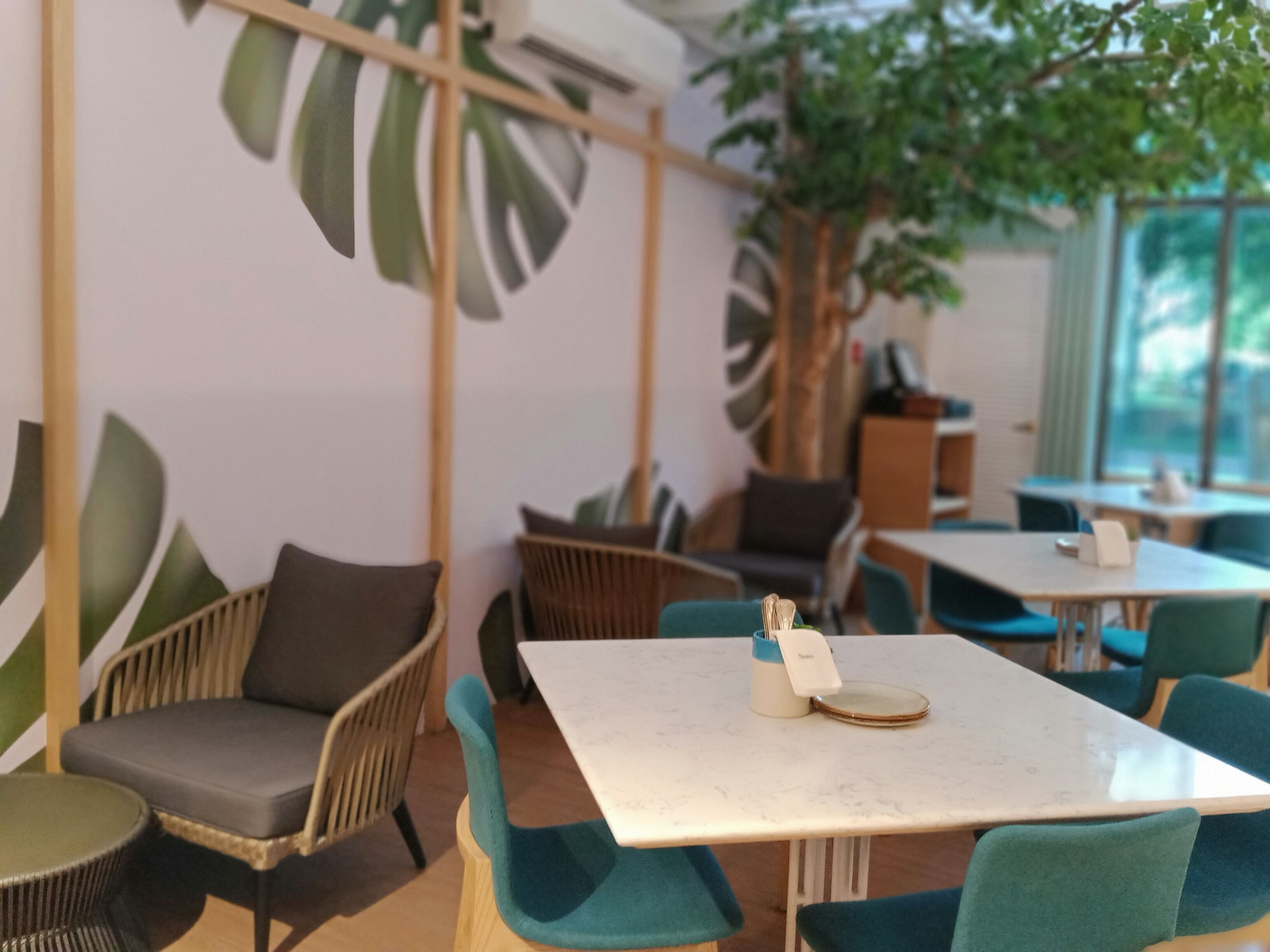 A modern cafe with wooden furniture, green chairs, and a large plant.