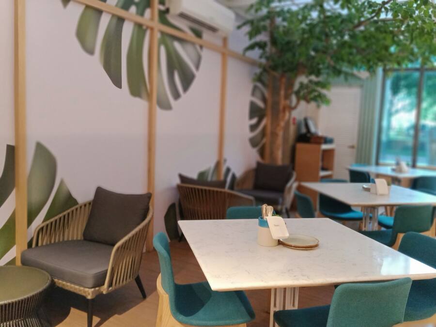 A modern cafe with wooden furniture, green chairs, and a large plant.