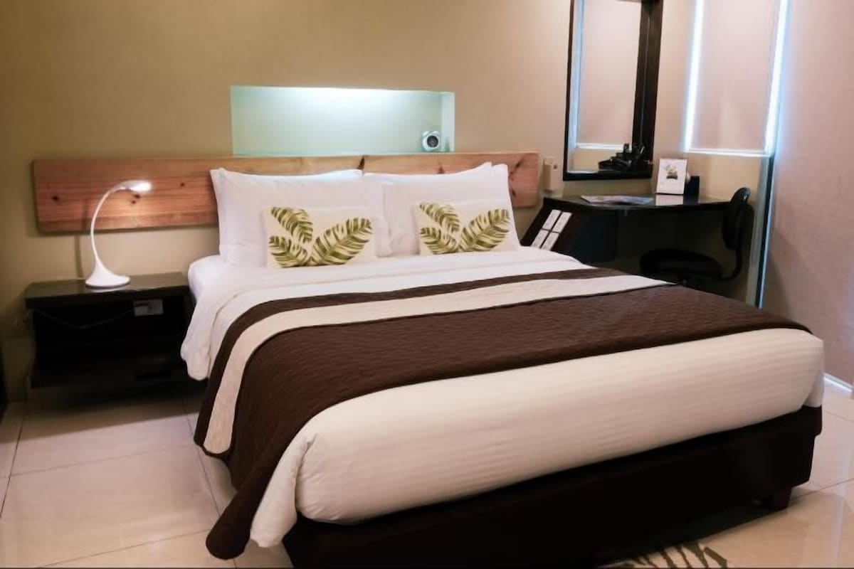 A hotel room with a bed, a nightstand, a desk, a lamp, a mirror, and a wall-mounted light fixture.