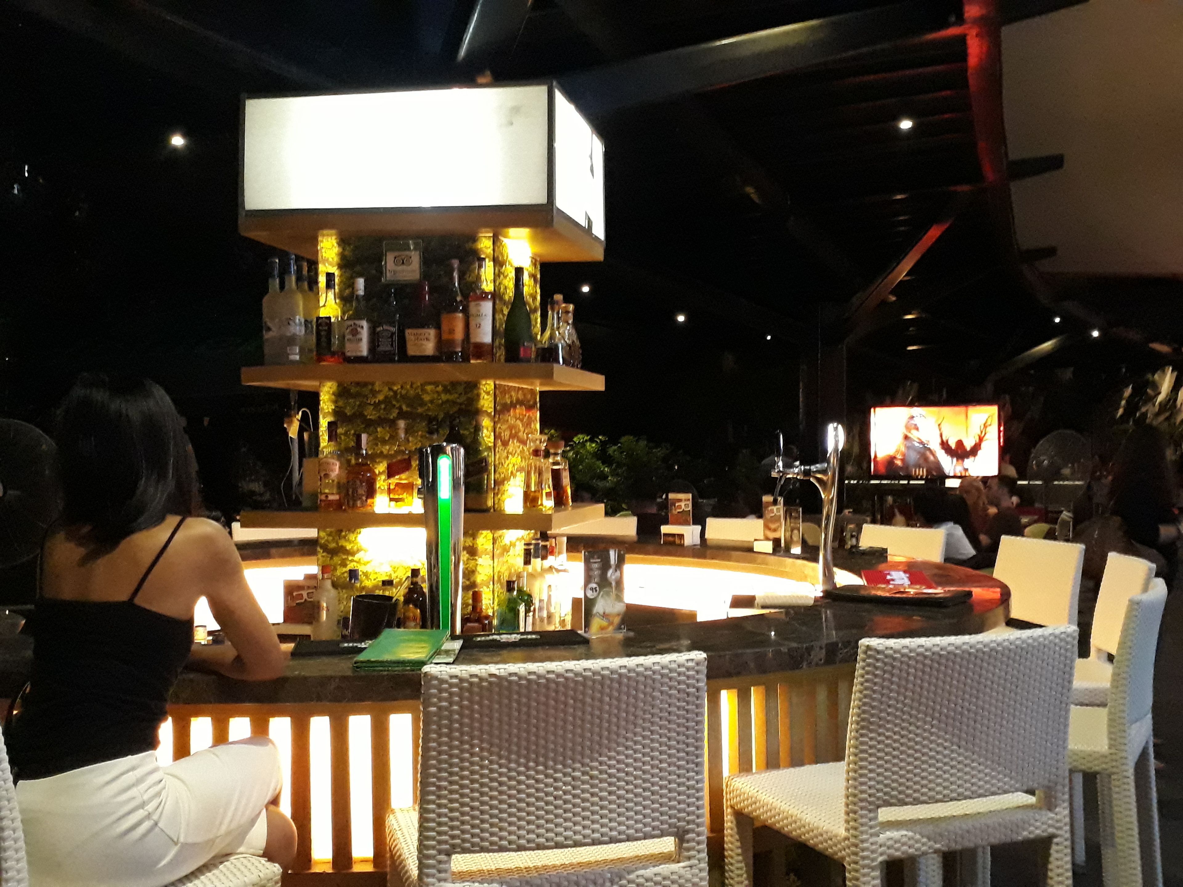 Zona de bar con estantería escalonada de botellas, una pantalla de televisión y sillas de mimbre.