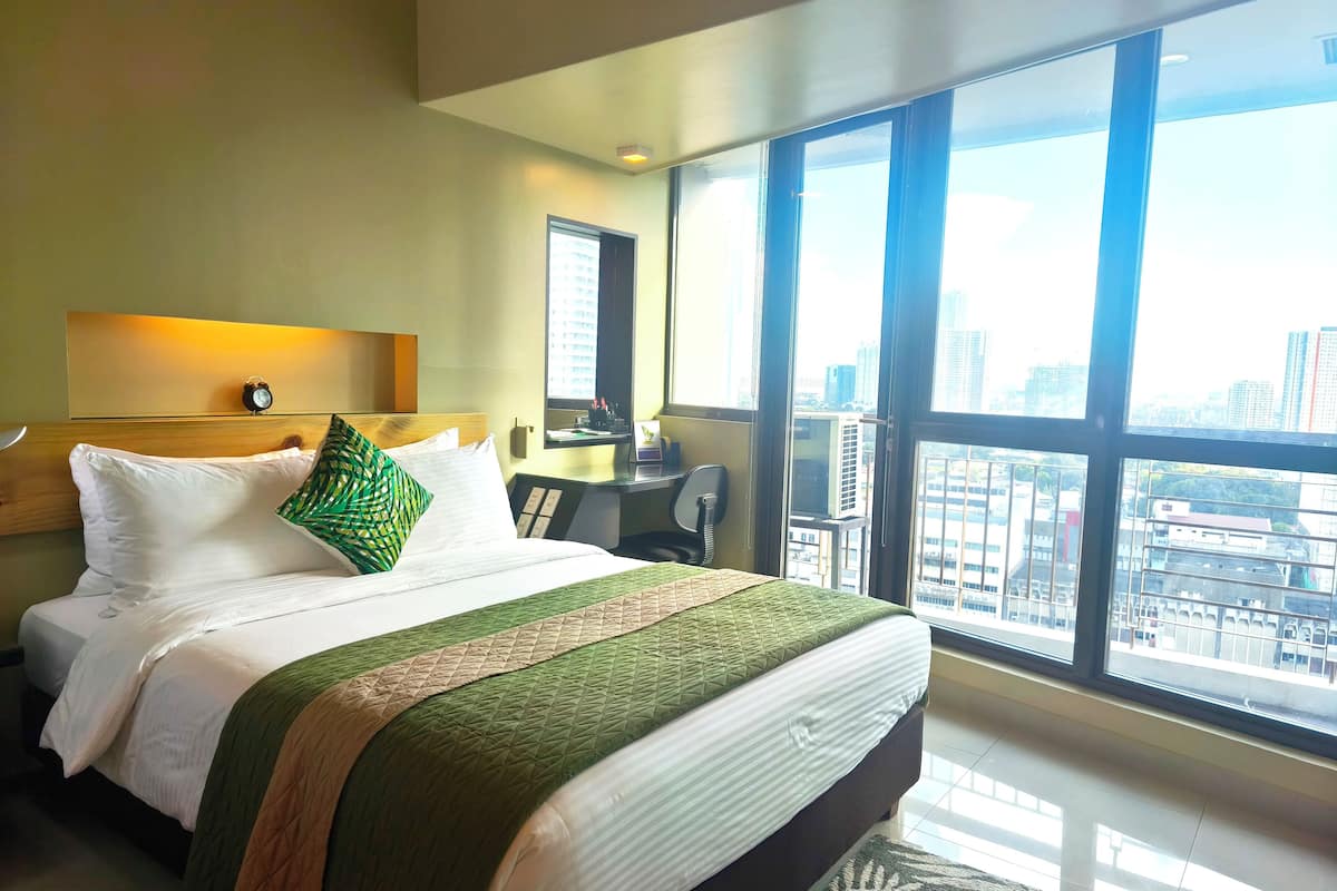 Quarto de hotel com uma cama grande, uma escrivaninha e vista da cidade através de amplas janelas.