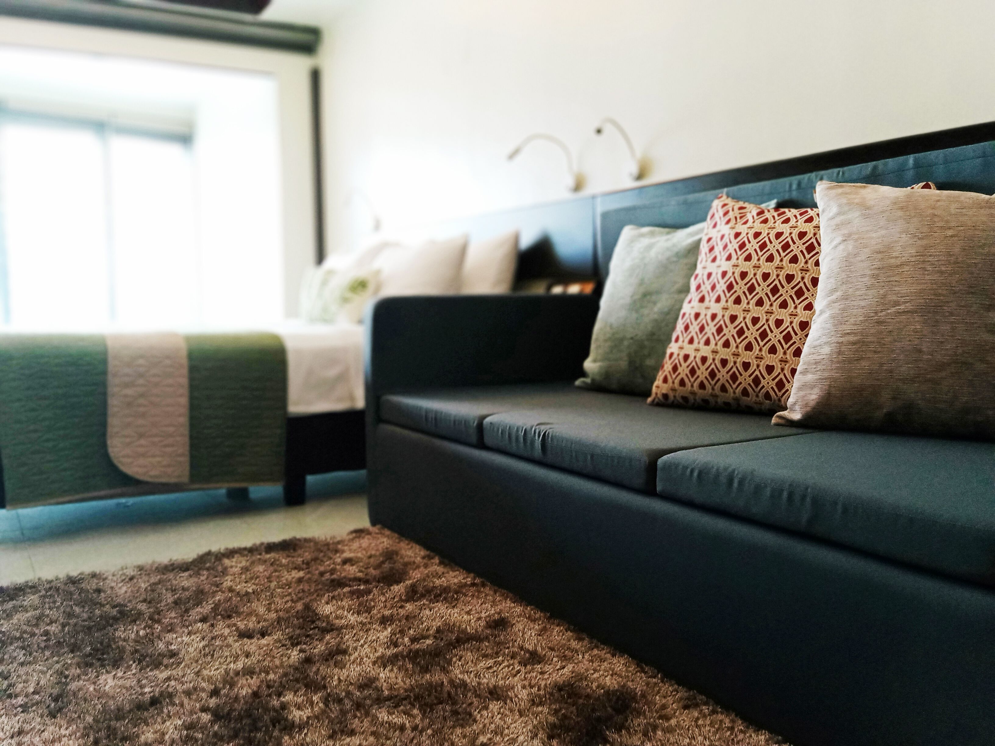 A modern living room with a dark sofa, multiple patterned cushions, a brown rug, and a bed with a green blanket.