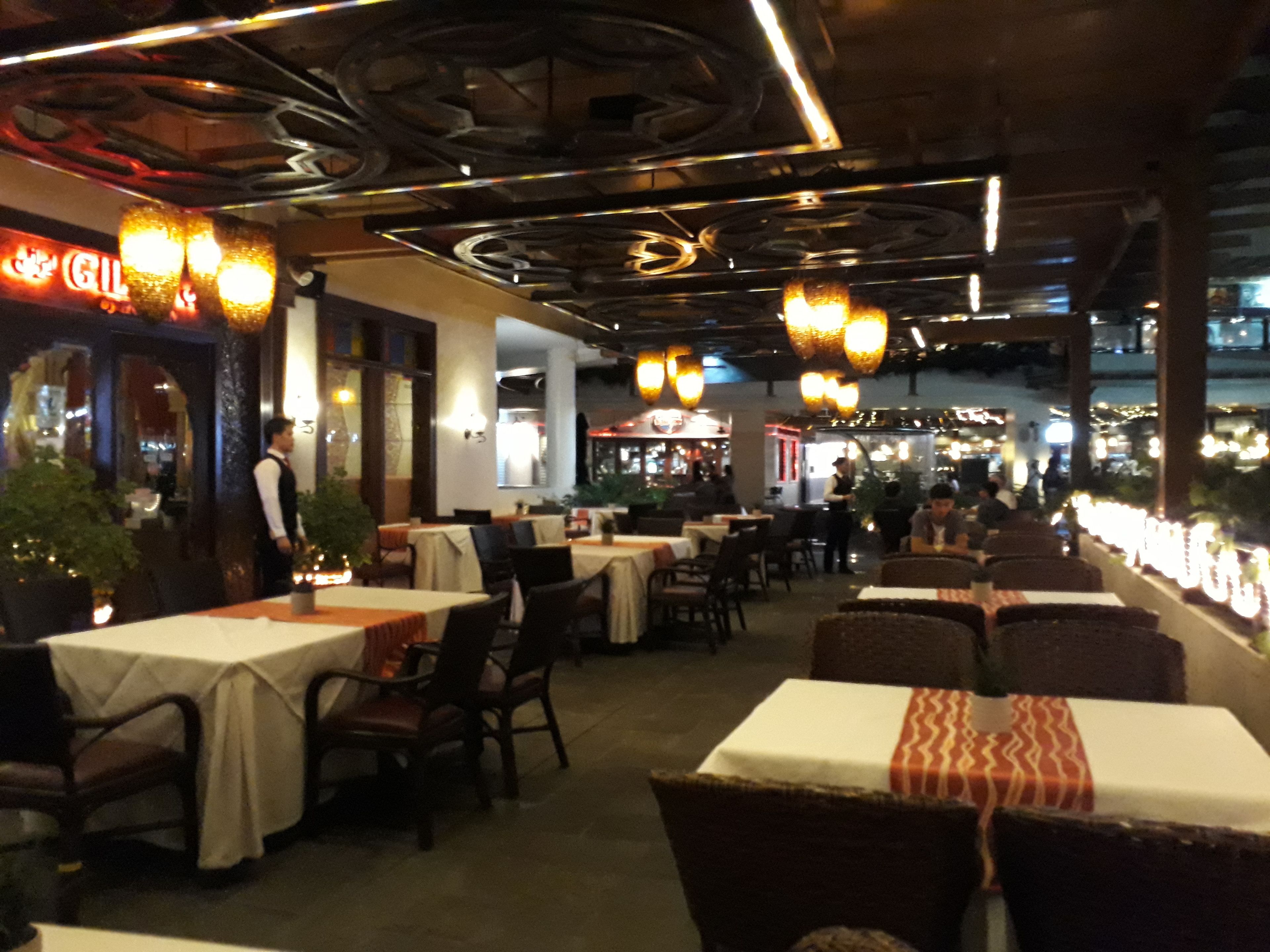 Un restaurante con mesas y sillas en el interior, luces colgantes y una zona de bar al fondo.