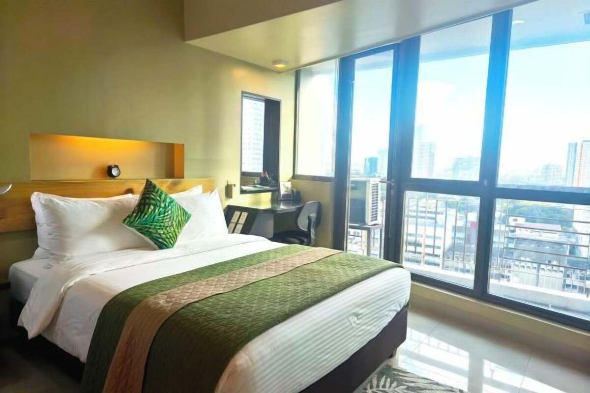 A hotel room with a large bed, a desk, a chair, and a view of the city through large windows.