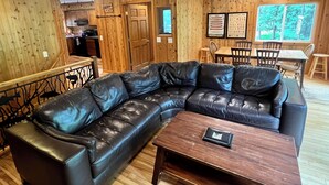 Un cuarto acogedor con un sofá seccional de cuero negro, una mesa de comedor de madera con sillas y una zona de cocina al fondo.