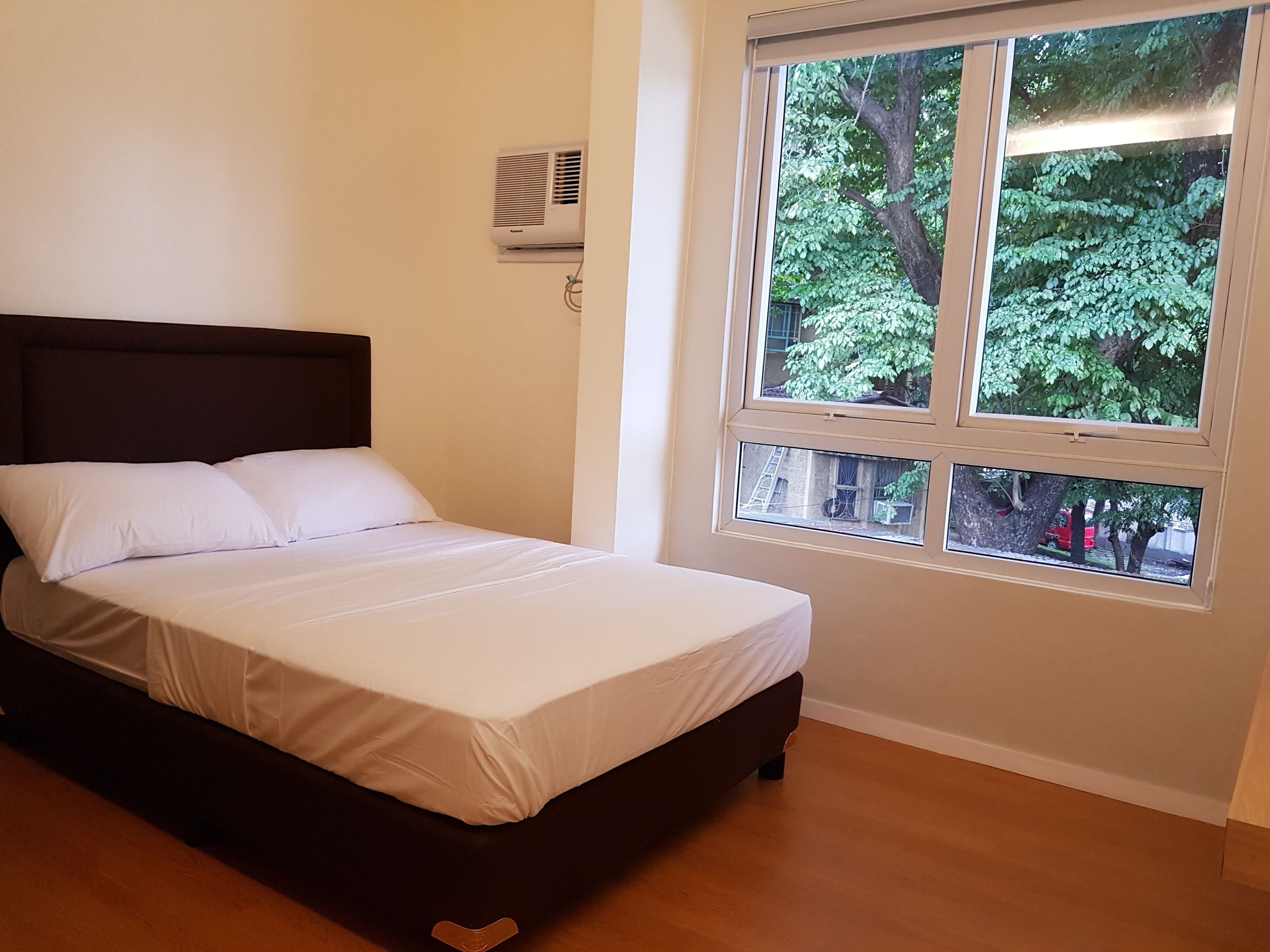 Una cama con ropa blanca en una habitación con ventana que da a un árbol.