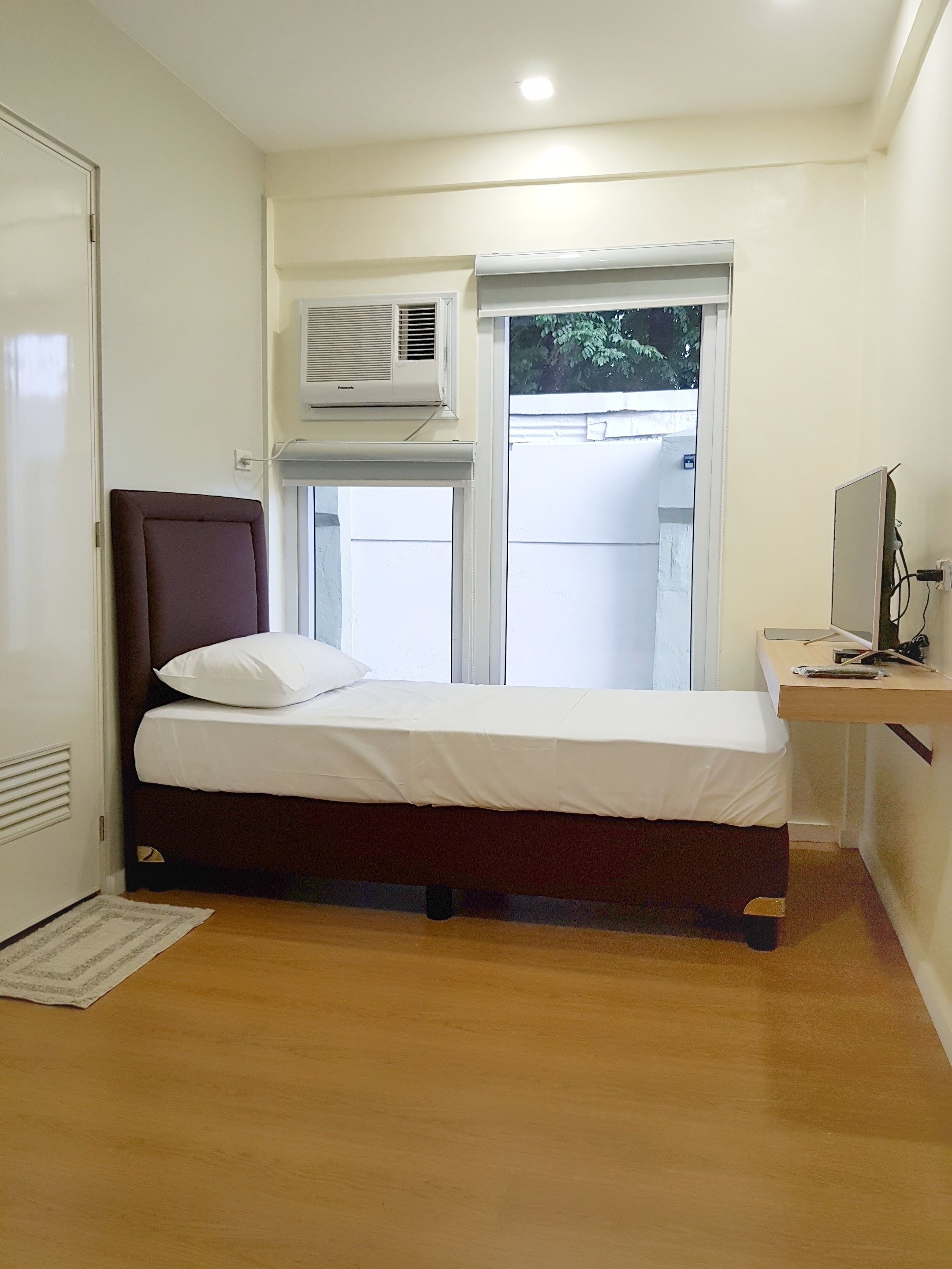 A bedroom with a bed, a desk, an air conditioner, and a window.