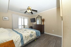 Un dormitorio con cama, cómoda, ventilador de techo, televisión y puerta que da a un balcón.