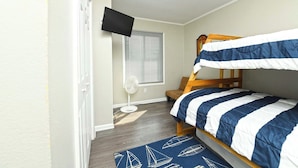 A bedroom with a bunk bed, a standing fan, a television on the wall, and a window with blinds.
