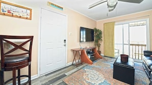 A living room with a ceiling fan, a TV, a plant, a patterned rug, and a door.