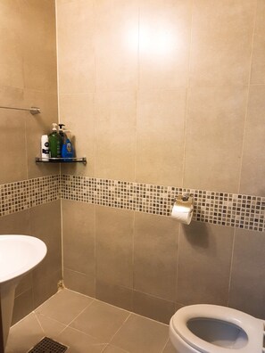 Un baño con baldosas color beige, un lavamanos blanco, un inodoro y un estante con artículos de tocador.