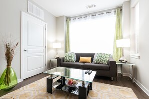 A modern living room with a glass coffee table, a sofa with patterned cushions, a side table with a lamp, and a vase with dried plants. - Montclair Apartments by Gracen Properties (Montclair)