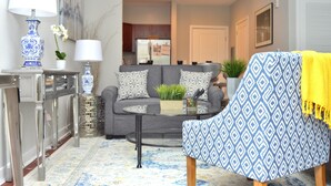 A living room with a grey sofa, a patterned armchair, a glass coffee table, and a decorative lamp. - Montclair Apartments by Gracen Properties (Montclair)