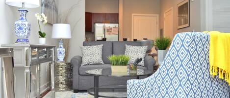 A living room with a grey sofa, a patterned armchair, a glass coffee table, and a decorative lamp.