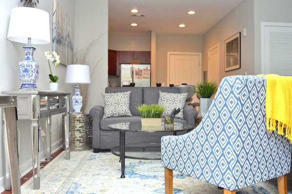 A living room with a grey sofa, a patterned armchair, a glass coffee table, and a decorative lamp.