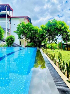 A swimming pool with a clear blue water surface, surrounded by lush green trees and a white building with a red roof.