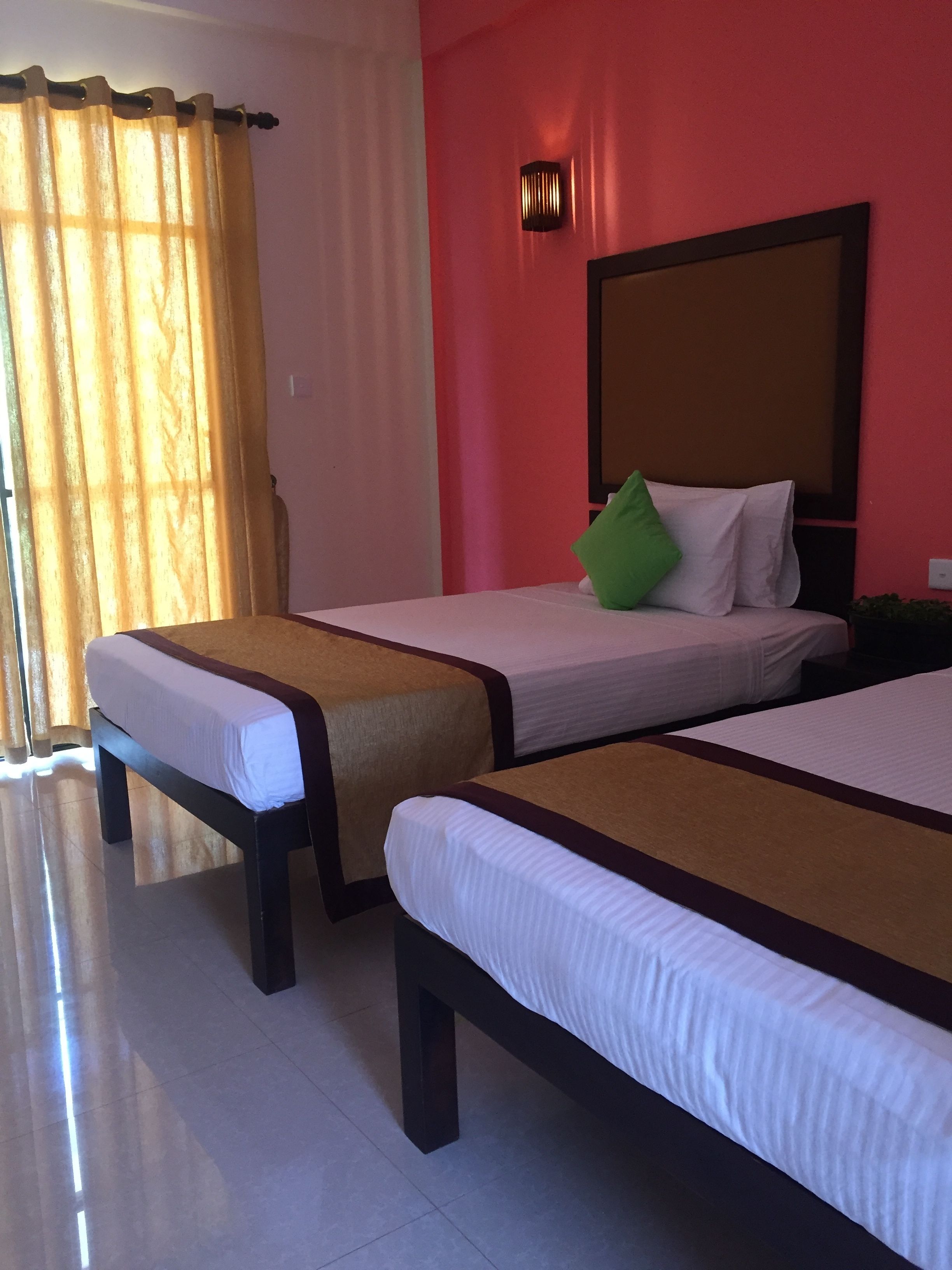 Habitación de hotel con dos camas individuales, un ventanal con cortinas, una lámpara de pared y una pared decorada en rojo.
