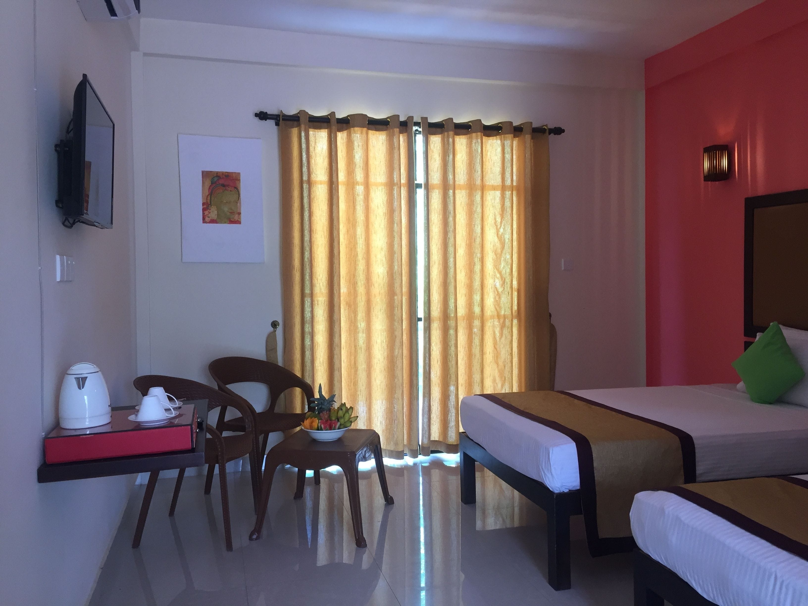 Une chambre d’hôtel avec un lit, un bureau avec une machine à café, une chaise, une petite table avec des fruits et une fenêtre avec des rideaux.