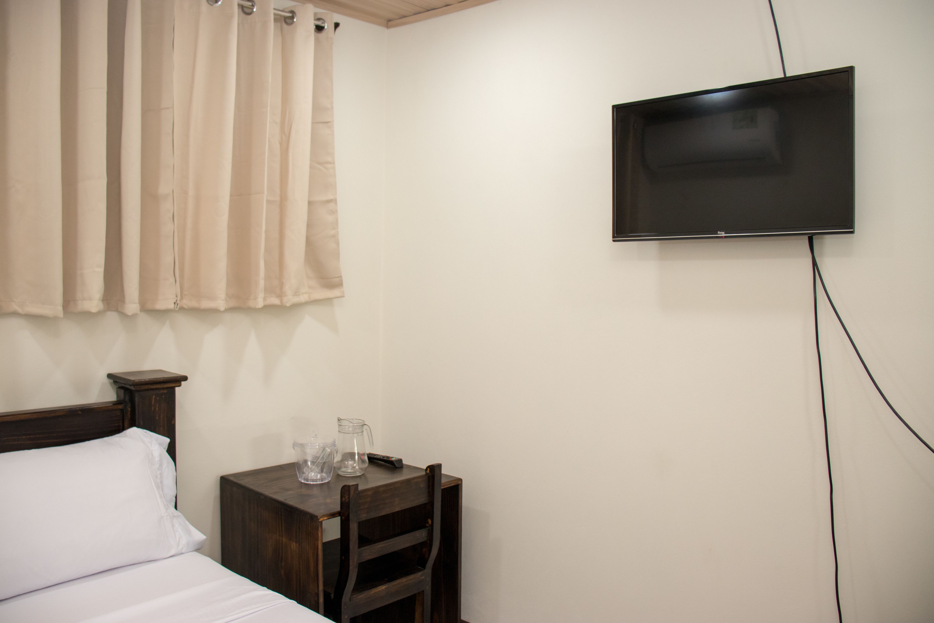 A hotel room with a bed, a wooden nightstand, a television mounted on the wall, and a window with curtains.