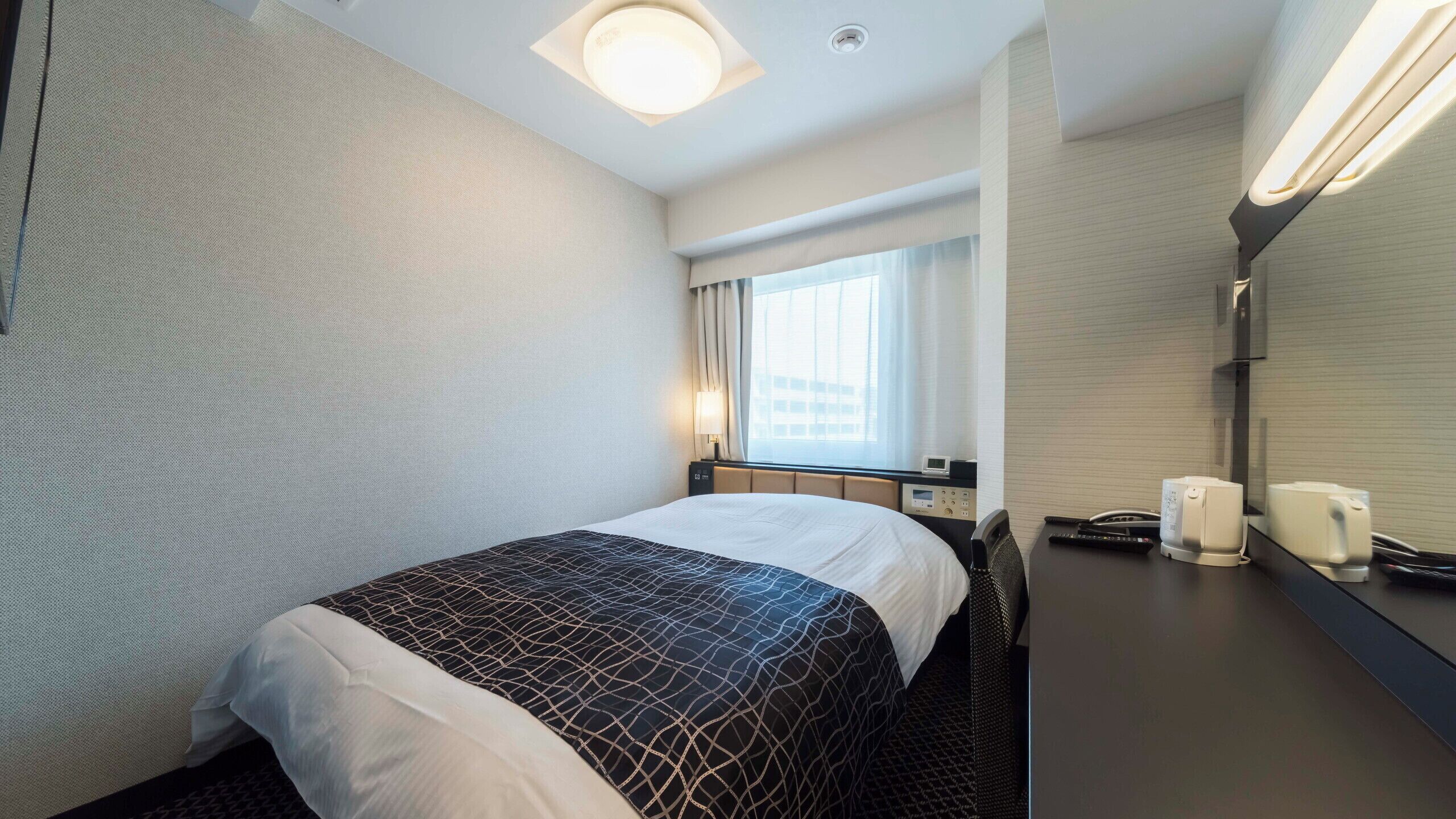 A hotel room with a bed, a desk with a phone and a coffee maker, a window with curtains, and a wall-mounted light fixture.