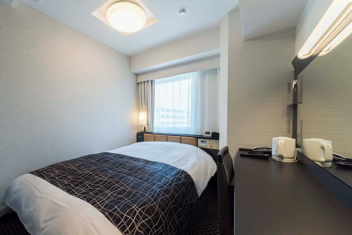 Habitación de hotel con cama, escritorio con teléfono y cafetera, ventana con cortinas, y lámpara de pared.