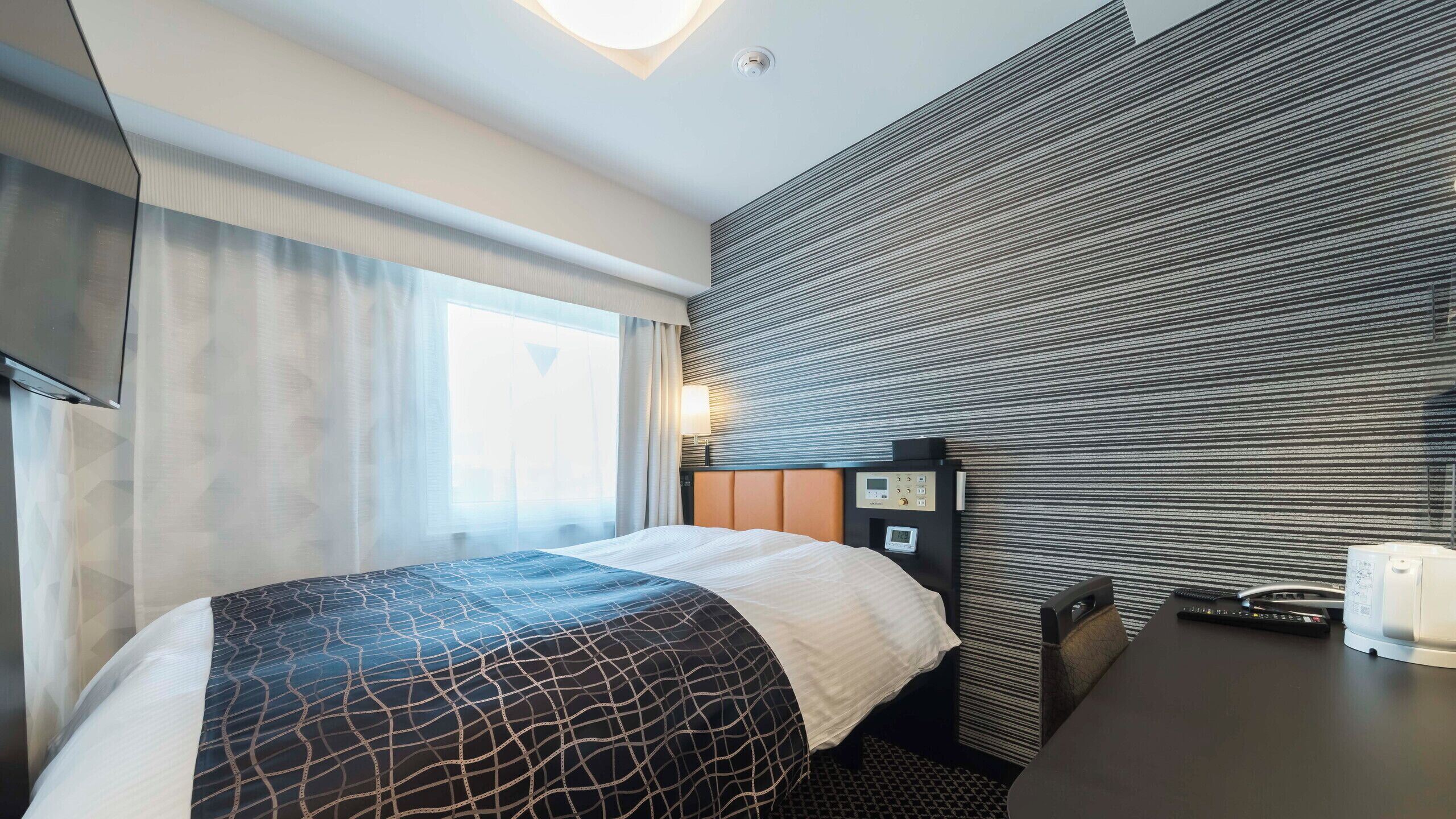 A hotel room with a bed, a desk, a control panel, and a window with curtains.