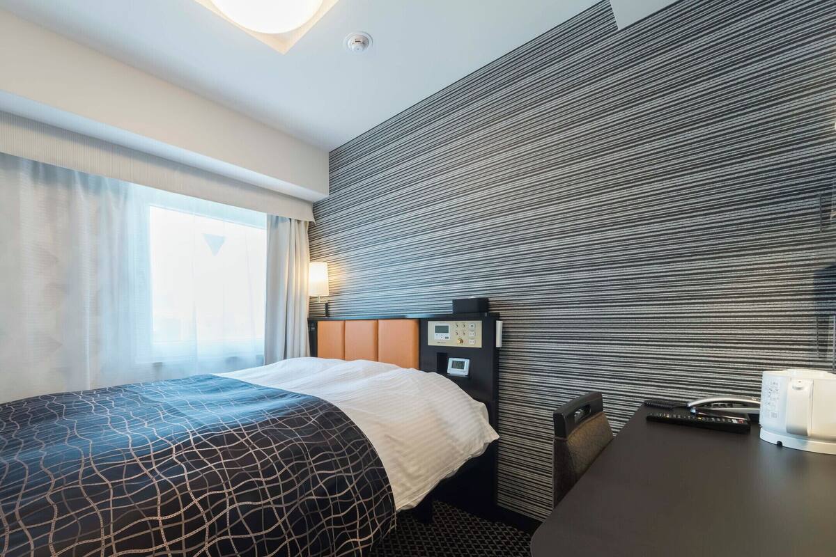 Una habitación de hotel con cama, escritorio, panel de control y ventana con cortinas.