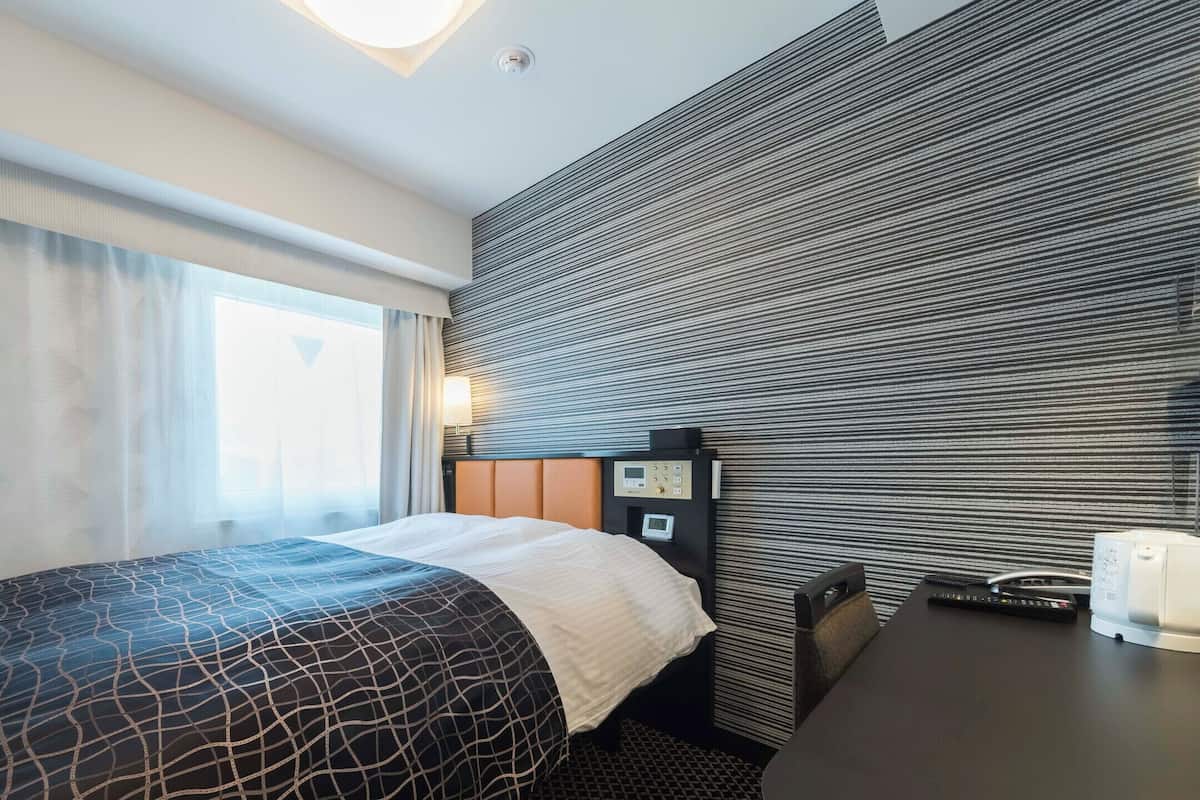 Hotellihuone, jossa on sänky, työpöytä, ohjauspaneeli ja ikkuna, jossa on verhot.