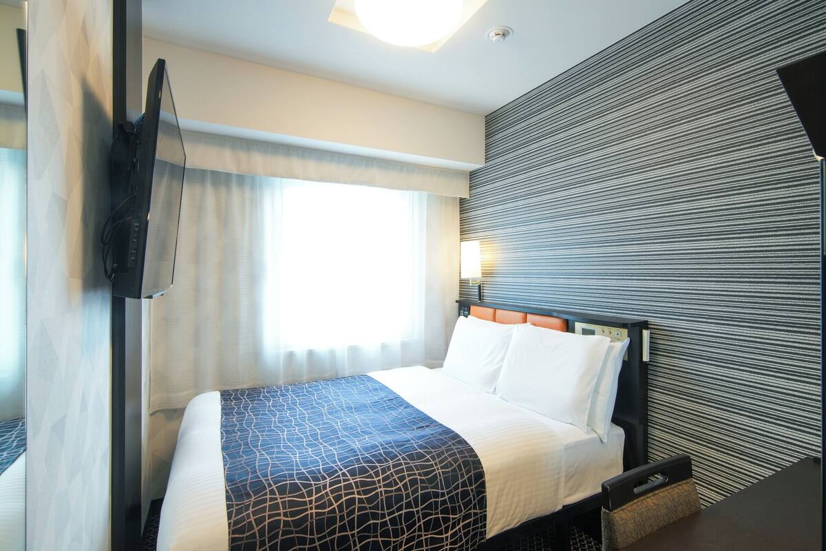Habitación de hotel con cama, televisión de pantalla plana montada en la pared, mesita de noche y ventana con cortinas.