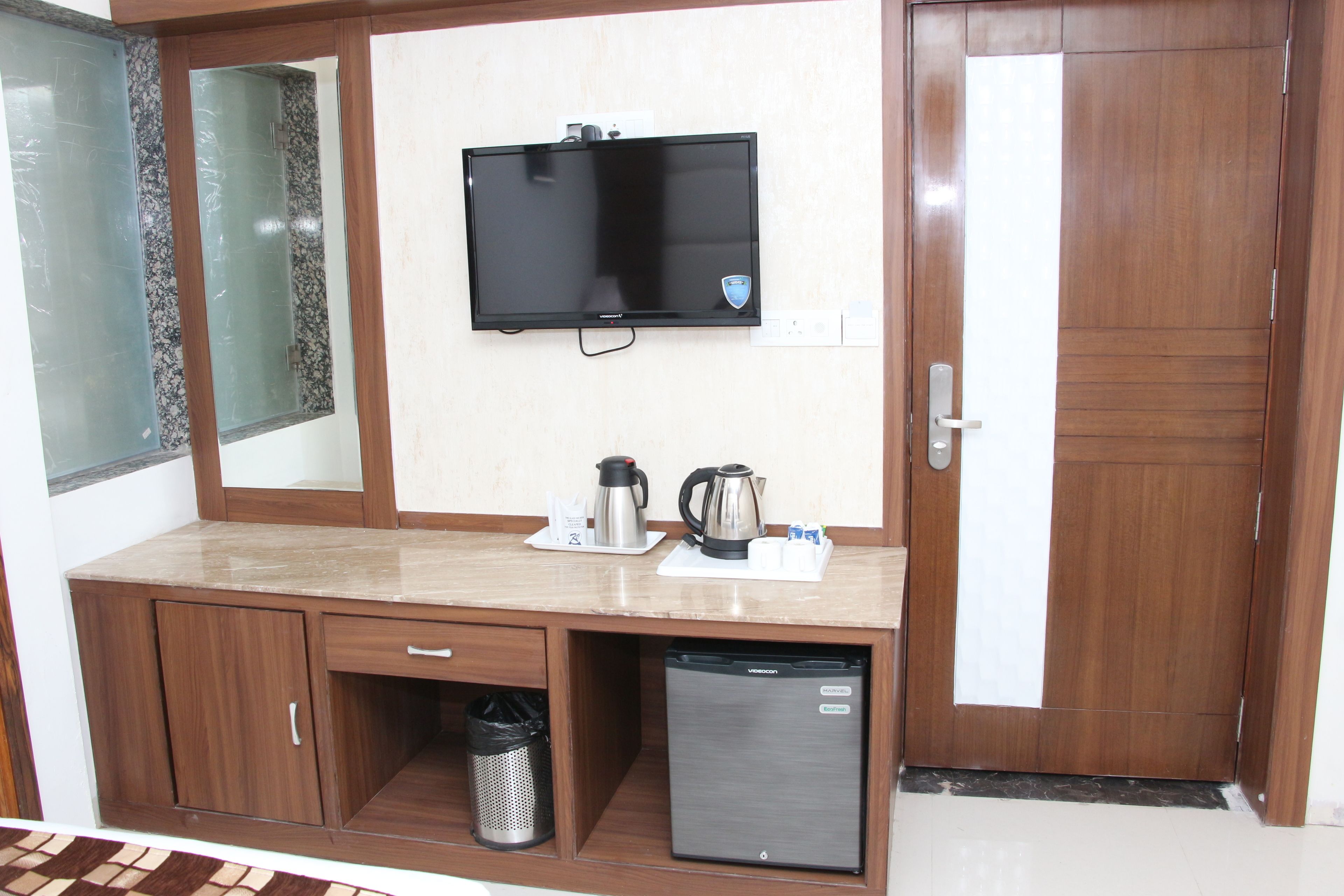 A hotel room with a wooden desk, a television mounted on the wall, a refrigerator, and a trash bin.