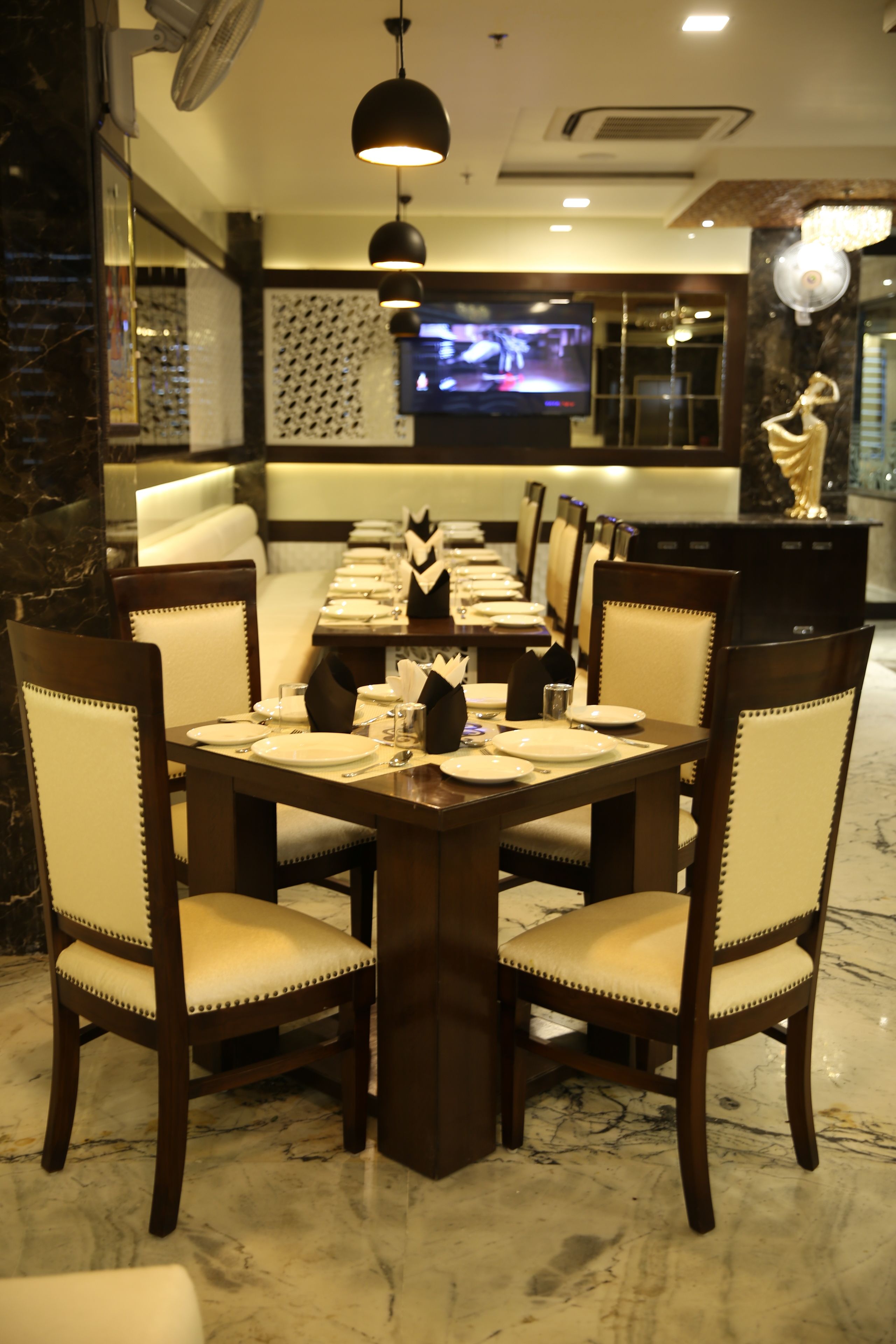 A dining area with a long table set for six, surrounded by upholstered chairs with a light fabric. A television is mounted on the wall in the background.