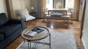 A cozy living room with a dark blue sofa, a round wooden coffee table with books, a white armchair, a small side table with a lamp, and a dining table with chairs.