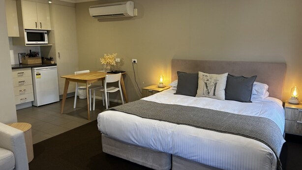 A bedroom with a large bed, bedside tables with lamps, a desk with a chair, and a kitchenette with a microwave and refrigerator.