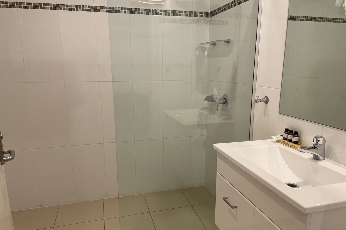 Un baño con cabina de ducha de vidrio, lavamanos blanco con espejo y estante con artículos de aseo.
