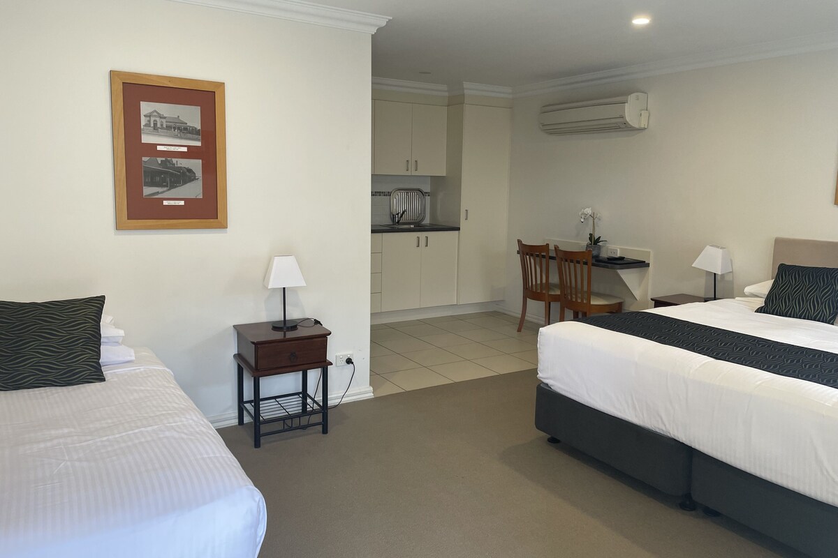 Habitación de hotel con dos camas, un escritorio, una silla, una mesita con lámpara y un cuadro en la pared.