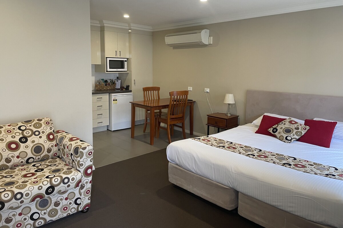 Quarto de hotel com cama, cozinha compacta, mesa de jantar com cadeiras e poltrona estofada com padrão.