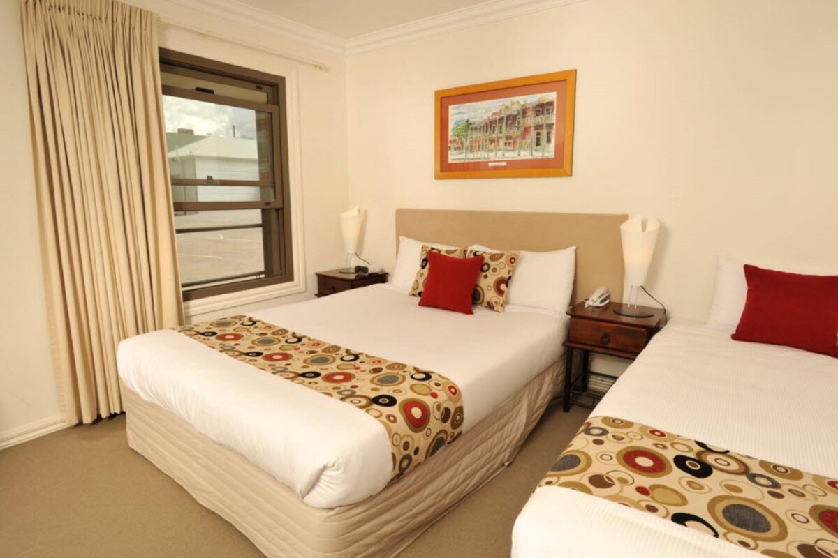 A hotel room with two beds, a window with curtains, a framed picture on the wall, and a bedside table with a lamp.