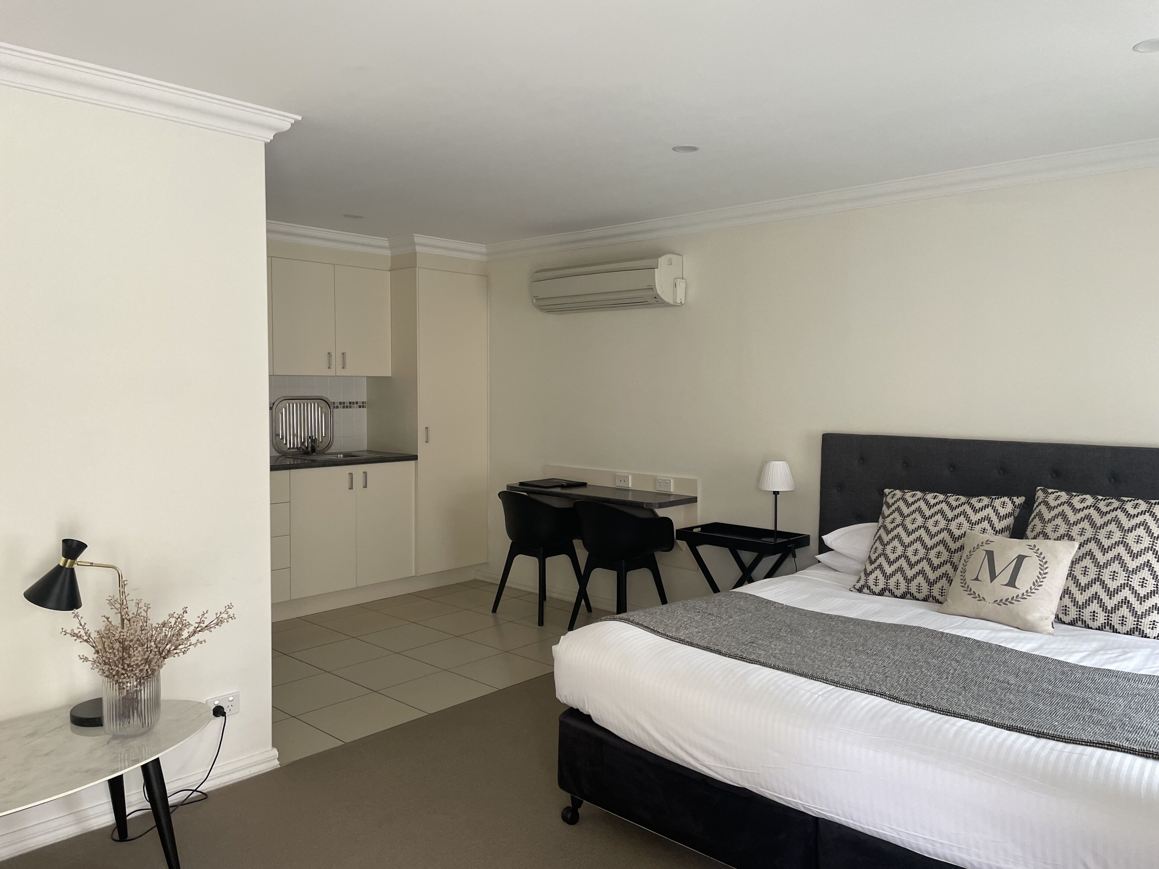 A bedroom with a bed, bedside table, lamp, and a view of a kitchen area.