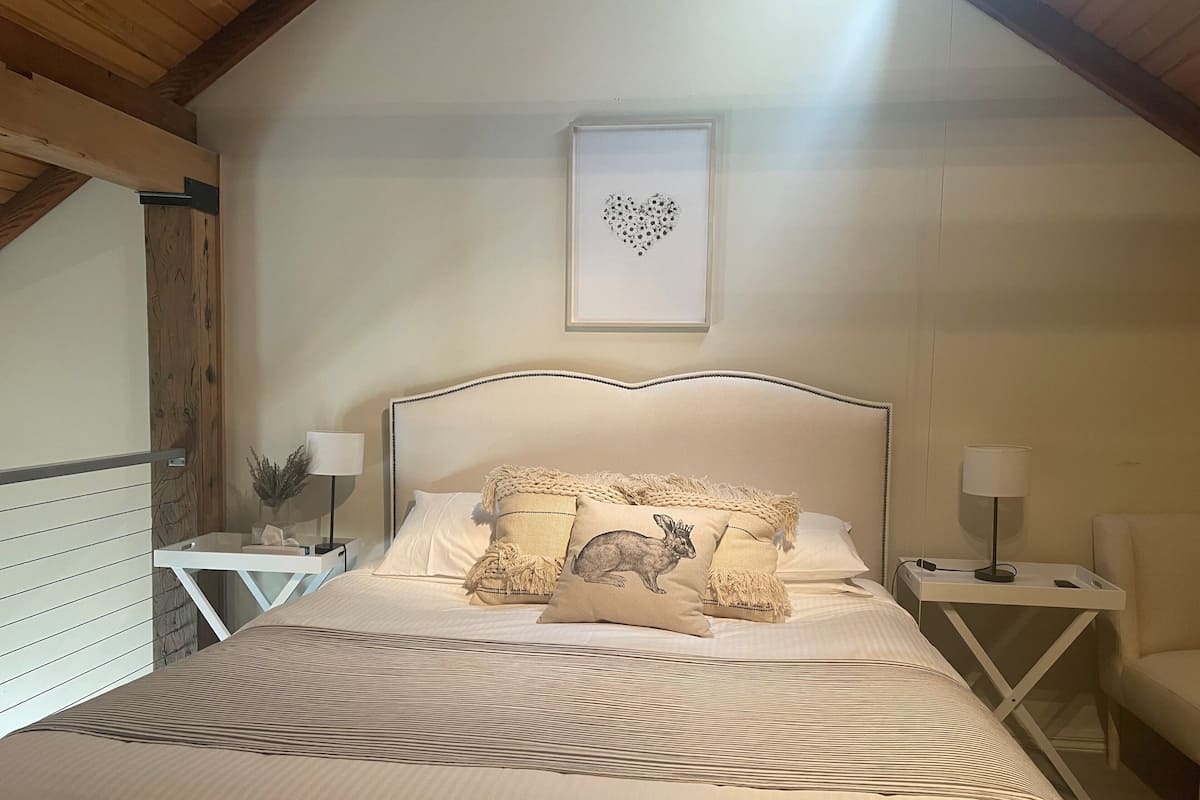 A bedroom with a bed, two bedside tables, a chair, a framed picture on the wall, and a wooden ceiling.