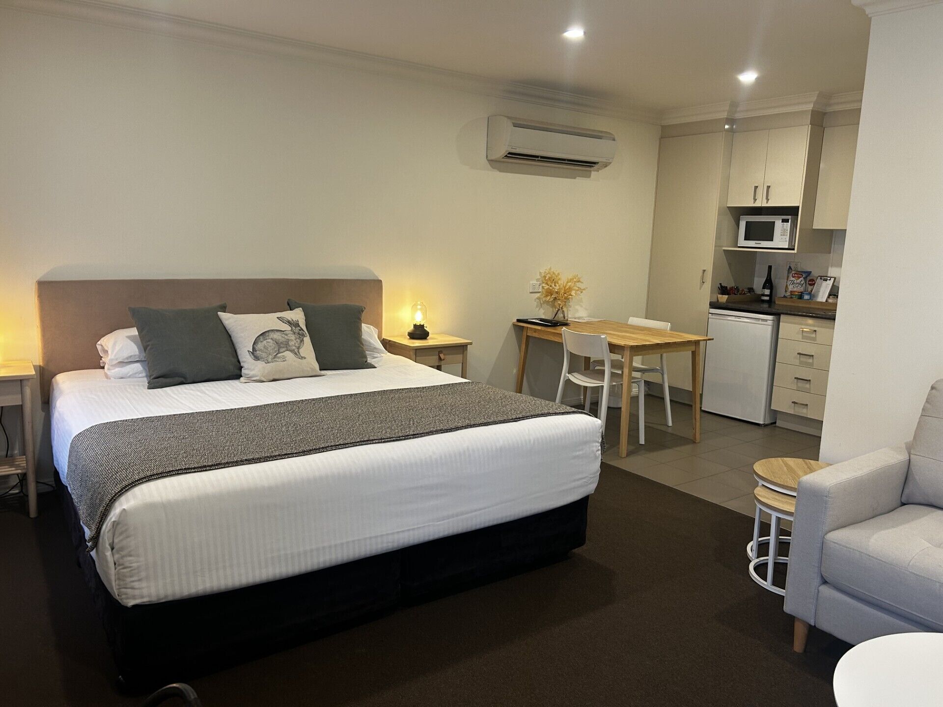 A hotel room with a bed, bedside table, lamp, desk, chair, kitchenette, and a sofa.