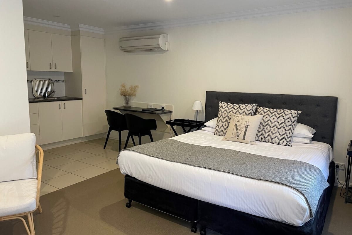 A bedroom with a bed, bedside table, and a desk with a chair. There is a kitchenette in the background.