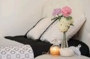 Ein Bett mit einem weiß-schwarz gemusterten Überwurf, zwei Kissen, einer Vase mit Blumen und einer kleinen Kerze auf einem Nachttisch.