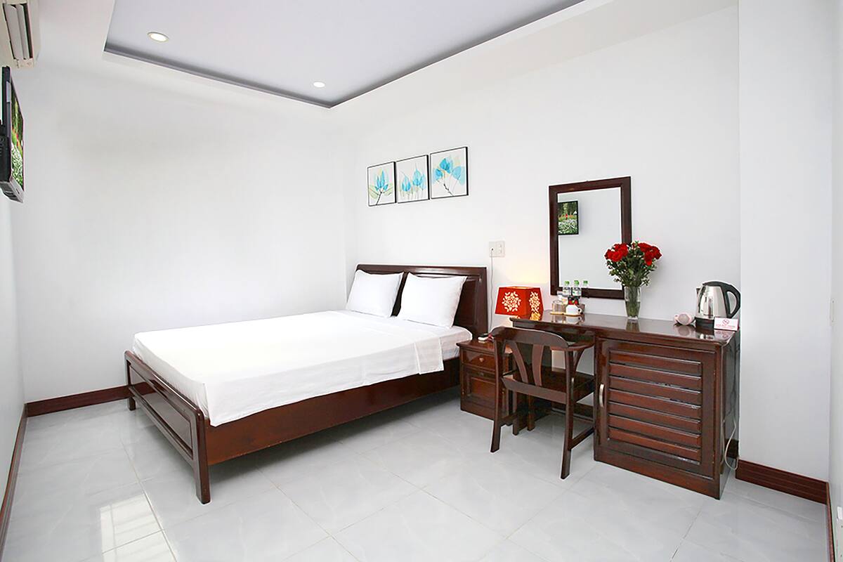 Quarto com cama, escrivaninha, cadeira, espelho e um vaso com flores.