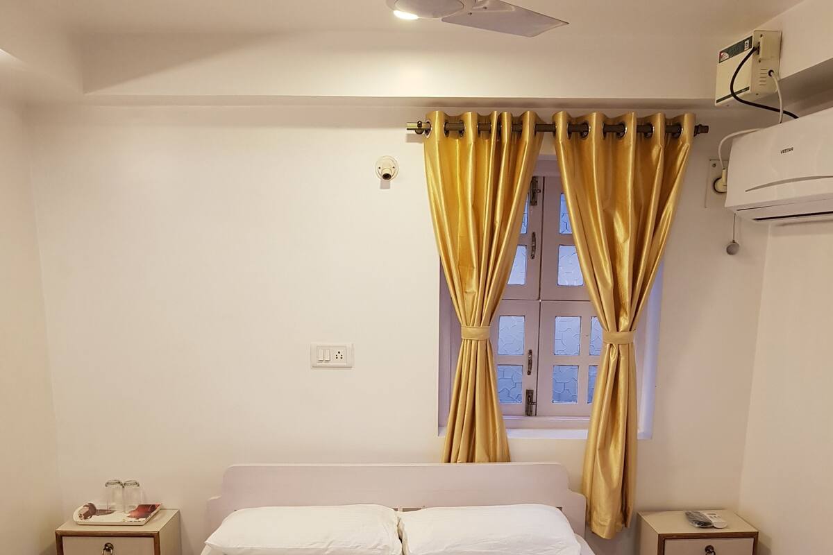 Una cama bien hecha con ropa blanca, una ventana con cortinas amarillas, un ventilador de techo y una mesita de noche con una lámpara.