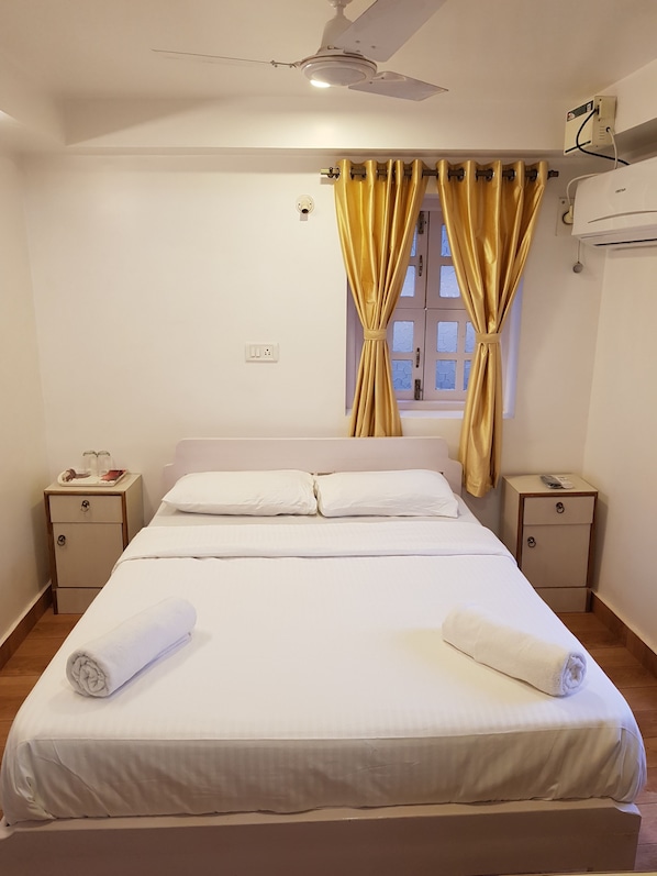 A neatly made bed with white linens, a window with yellow curtains, a ceiling fan, and a small bedside table with a lamp.