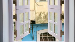 A window with white frames and open shutters, revealing a view of a building with a blue floor and a window with a metal grill. - Marquito's Guest House (Panaji)
