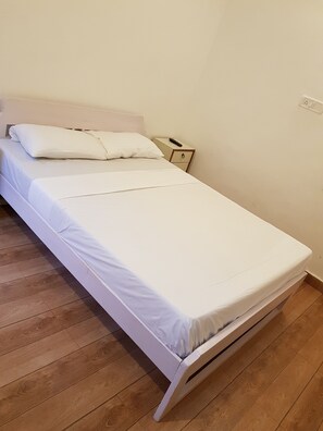 Ein Einzelbett mit weißer Bettwäsche, ein Nachttisch aus Holz mit Fernbedienung und eine schlichte Wand mit Steckdose.