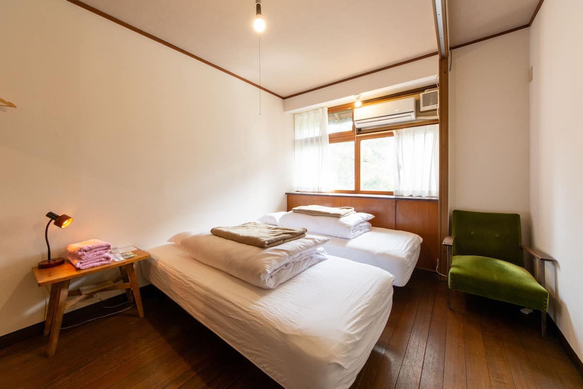 A room with two beds, a green chair, a bedside table with a lamp, and a window with curtains.