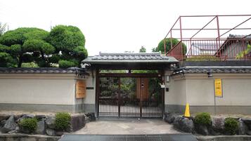 Un portón tradicional japonés con techo de tejas, un muro de piedra con arbustos pequeños y un letrero amarillo.